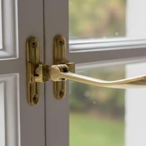 May include: Close-up of a white window frame with a brass handle. The handle is curved and attached to the frame with brass hardware. The window is divided into panes, with a blurred green background visible through the glass.