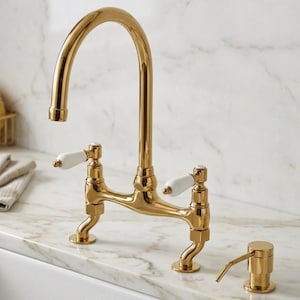 May include: A polished gold kitchen faucet with a high-arch spout and two white ceramic lever handles. The faucet features a bridge-style design and is accompanied by a separate matching gold liquid soap dispenser pump.