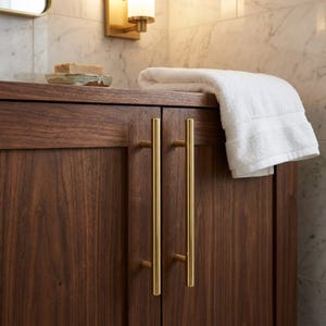May include: A dark brown wooden bathroom vanity cabinet featuring two vertical brushed gold metal bar handles. A folded white cotton hand towel rests on the top surface next to a small rectangular bar of soap on a ceramic dish.