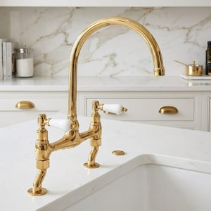 Peut inclure: Un robinet de cuisine en or poli avec un bec haut et deux poignées à levier blanches. Le robinet présente un design de style pont avec deux supports de base séparés, un col incurvé et une finition métallique, installé sur un comptoir blanc.