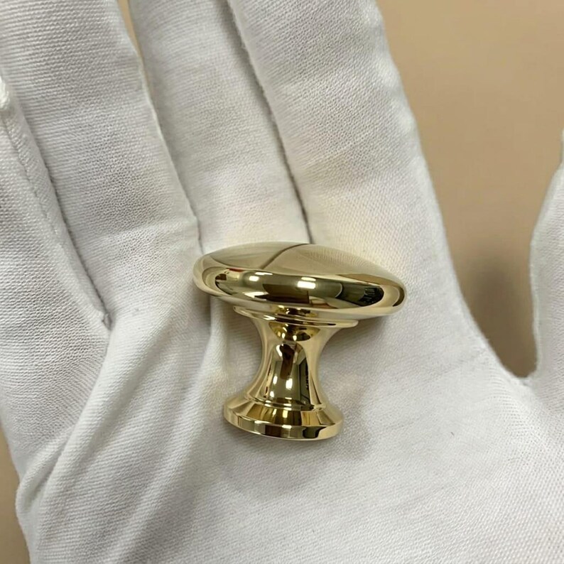 May include: A polished gold-tone metal cabinet knob featuring a smooth, rounded top surface and a tapered, flared base. The hardware has a reflective, high-gloss finish and a circular profile, designed for attachment to furniture or cabinetry doors.
