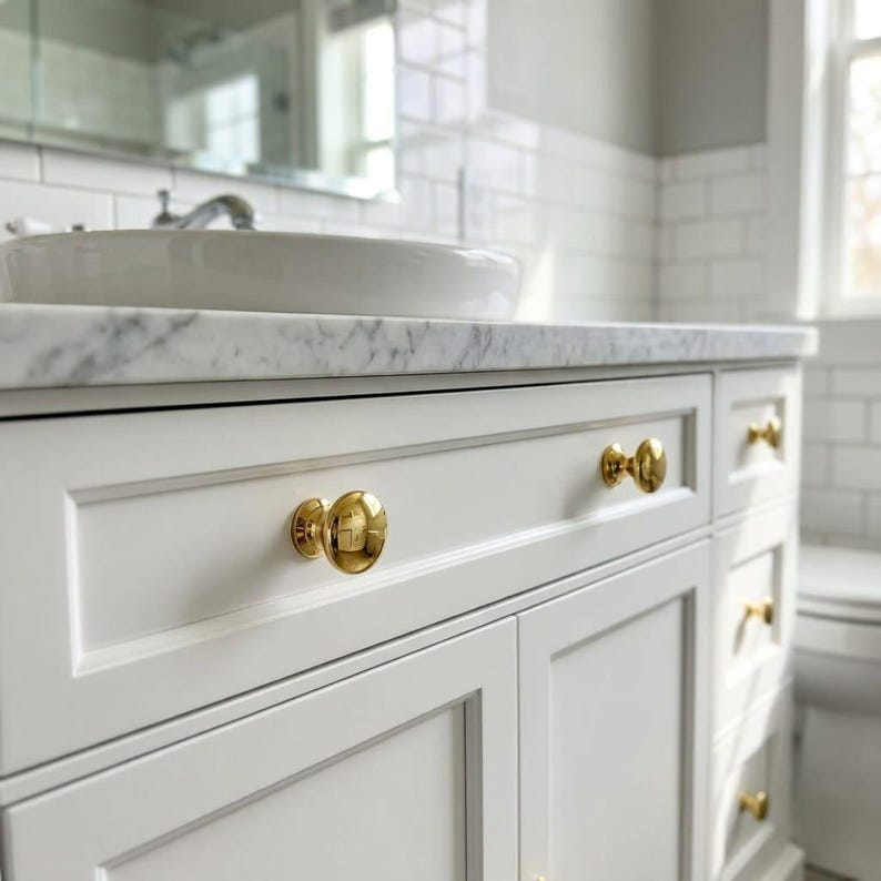 May include: A white bathroom vanity cabinet featuring a marble countertop and a white ceramic sink basin. The cabinet includes multiple drawers and doors, each accented with polished gold-toned round knobs, providing a bright and clean aesthetic.