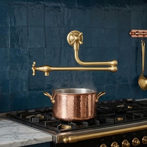 Könnte beinhalten: Ein an der Wand montierter Messing-Topffüller mit doppeltem Schwenkarm und Kreuzgriff-Design, positioniert über einem gehämmerten Kupfertopf auf einem schwarzen Gasherd mit einer sichtbaren Marmorarbeitsplatte.