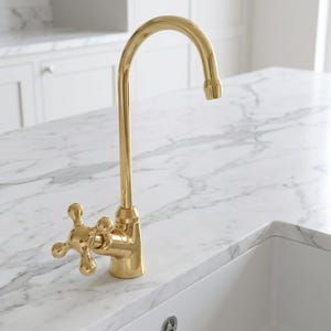 May include: A polished gold kitchen faucet featuring a high-arc spout and a single cross-style handle. The faucet has a smooth, reflective metallic finish and a classic design with a curved neck, positioned on a white marble countertop surface.