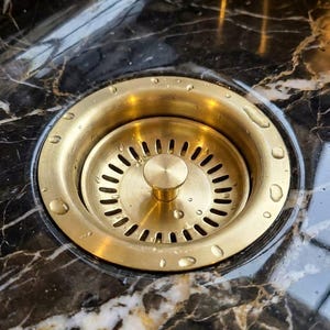 May include: A circular brushed brass kitchen sink drain strainer with a central knob and a series of radial slots. The metallic gold-toned fixture is set into a dark black marble countertop featuring prominent white and gold veining with water droplets.