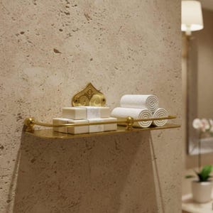 May include: A gold-toned bathroom shelf with rolled white towels and wrapped soaps. The shelf is mounted on a textured beige wall, with a decorative gold bracket above. A wall sconce and a potted plant are visible.