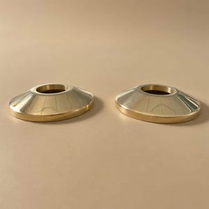 May include: Two gold-colored candle holders with a wide, flat base and a conical top. The candle holders have a shiny, reflective surface and a dark brown interior. The bases are a slightly darker shade of gold.