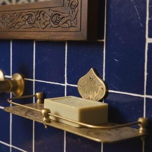 May include: A brass-toned soap shelf with a bar of soap, mounted on blue tiled wall. The shelf has an ornate design, and a decorative brass accent is above it. A wooden framed mirror is above the tiles.
