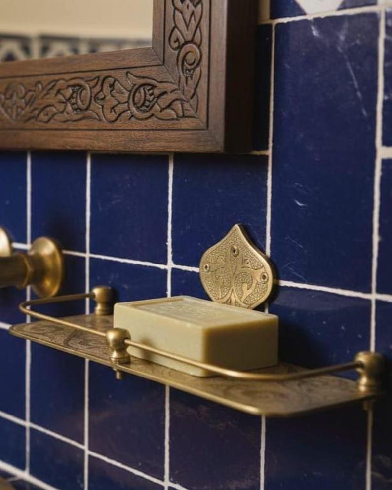May include: A brass-toned soap shelf with a bar of soap, mounted on blue tiled wall. The shelf has an ornate design, and a decorative brass accent is above it. A wooden framed mirror is above the tiles.