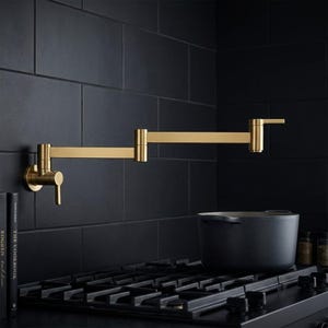 Può includere: Un rubinetto riempitore per pentole a parete con finitura in oro spazzolato e braccio oscillante a doppio snodo. Il dispositivo presenta una singola maniglia a leva alla base e una maniglia secondaria sul beccuccio, montato su una parete di piastrelle scure.