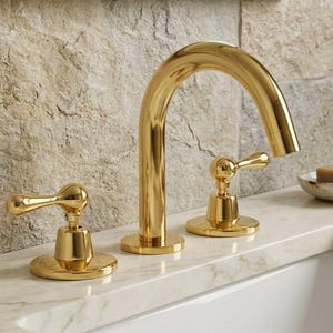 Könnte beinhalten: Ein poliertes goldenes Badezimmerarmaturen-Set mit einem hoch geschwungenen Auslauf und zwei separaten Hebelgriffen. Die Armatur hat eine reflektierende metallische Oberfläche und ist auf einer weißen Marmorarbeitsplatte montiert.
