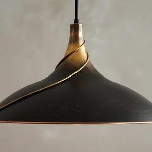 Puede incluir: Una lámpara colgante negra con la parte superior dorada y un borde cobrizo. Un alambre dorado en espiral rodea la parte superior de la lámpara. La lámpara cuelga de un cable negro. La lámpara tiene un diseño moderno e industrial.