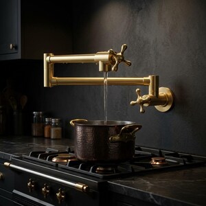 May include: A wall-mounted, brushed gold pot filler faucet with two cross-style handles and an articulated swing arm. Water flows from the spout into a dark, hammered metal pot resting on a black gas stovetop with multiple burners and knobs.