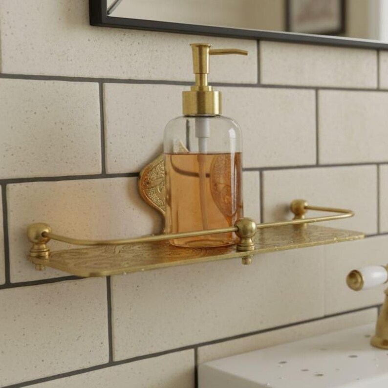 May include: A clear glass soap dispenser with a gold pump sits on a gold-colored shelf. The shelf is mounted on a white tiled wall. The dispenser is filled with a light orange liquid.