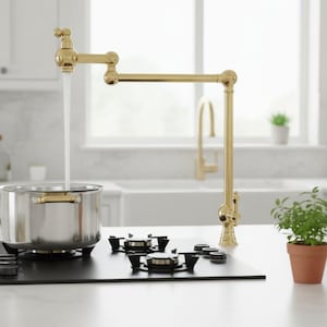 Puede incluir: Un grifo dorado para llenar ollas se extiende sobre una olla de acero inoxidable en una encimera negra. El agua fluye del grifo a la olla. Una pequeña planta en maceta y una botella de vidrio están en la encimera.