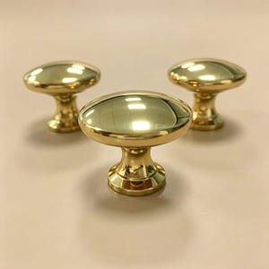 May include: Three polished brass cabinet knobs with a smooth, rounded top and a tapered base. Each knob features a reflective, metallic gold-toned finish with a circular head and a sturdy stem, arranged in a triangular formation on a plain surface.