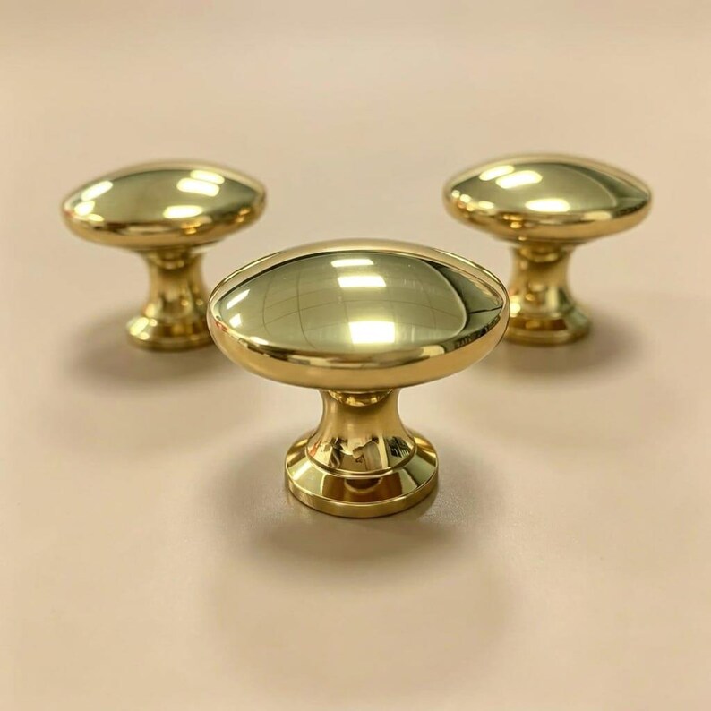 May include: Three polished brass cabinet knobs with a smooth, rounded top and a tapered base. Each knob features a reflective, metallic gold-toned finish with a circular head and a sturdy stem, arranged in a triangular formation on a plain surface.