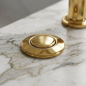 Op de afbeelding: Een close-up van een goudkleurige, ronde knop met een verhoogd midden, waarschijnlijk een onderdeel van een wastafel of kraan. Het ligt op een marmeren oppervlak met waterdruppels, naast een gouden kraanvoet en een parfumflesje.