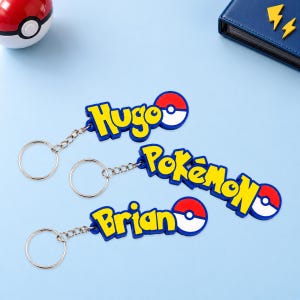 Puede incluir: Tres llaveros de goma flexible con letras amarillas de borde azul y un emblema circular rojo y blanco. Dos llaveros muestran los nombres Hugo y Brian, mientras que el tercero presenta el texto Pokémon, todos unidos a anillas de metal.