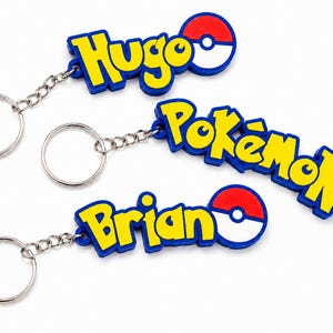 Könnte beinhalten: Drei Schlüsselanhänger mit gelber Schrift auf blauem Grund und einem rot-weißen Pokéball-Symbol. Ein Anhänger zeigt den Namen Hugo, einer Pokémon und einer Brian. Jeder Anhänger ist mit einer kurzen Kette an einem silbernen Metallring befestigt.