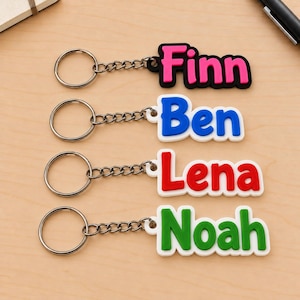 Könnte beinhalten: Vier personalisierte Schlüsselanhänger mit Metallringen und Ketten. Jeder zeigt einen Namen in fetter, erhabener Schrift auf weißem oder schwarzem Grund: Finn in Pink auf Schwarz, Ben in Blau auf Weiß, Lena in Rot auf Weiß und Noah in Grün auf Weiß.