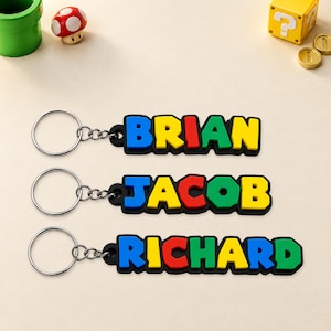 Pode incluir: Três porta-chaves com os nomes BRIAN, JACOB e RICHARD em letras coloridas, grossas e multicoloridas. Cada nome está preso a um anel de metal prateado por uma corrente curta, sobre um fundo de cor clara.