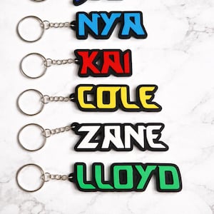 Könnte beinhalten: Schlüsselanhänger mit den Namen: Jay, Nya, Kai, Cole, Zane und Lloyd. Jeder Schlüsselanhänger zeigt einen anderen Namen in fetter, farbiger Schrift mit schwarzem Rand. Die Schlüsselanhänger sind an silbernen Schlüsselringen befestigt.