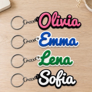 Könnte beinhalten: Vier personalisierte Schlüsselanhänger mit Metallringen und Ketten. Jeder zeigt einen Namen in erhabener Schreibschrift: Olivia in Pink auf Schwarz, Emma in Blau auf Weiß, Lena in Grün auf Weiß und Sofia in Weiß auf Schwarz.