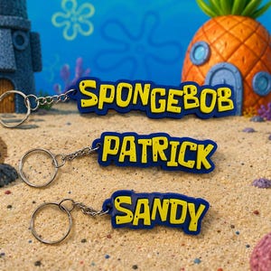 Op de afbeelding: Drie sleutelhangers met de namen SpongeBob, Patrick en Sandy in gele blokletters op een donkerblauwe achtergrond. Elke sleutelhanger bevat een zilverkleurige metalen ring bevestigd aan een korte ketting, met de namen duidelijk zichtbaar.