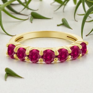 Op de afbeelding: Een gouden ring met zeven ronde, helderrode robijnen. De ring heeft een klassiek ontwerp, waarbij elke robijn stevig wordt vastgehouden. De achtergrond is zacht en neutraal met groene sprietjes.