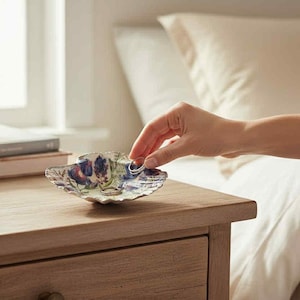 Könnte beinhalten: Eine kleine, dekorative Keramikschale mit gewelltem Rand, mit einem Blumendesign in Blau-, Lila- und Grüntönen. Die Schale steht auf einem Nachttisch aus Holz, mit einer Hand, die sich danach ausstreckt. Eine Sukkulente im Topf und Bücher sind ebenfalls vorhanden.