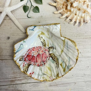 Puede incluir: Concha decorativa pintada con un diseño de tortuga marina roja y marrón. La concha tiene un borde dorado y está colocada sobre un fondo de una estrella de mar, otra concha y vegetación. La tortuga está rodeada de algas marinas.