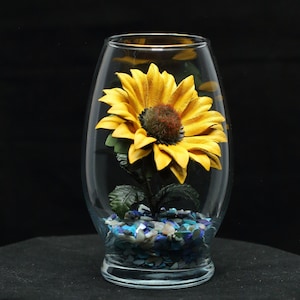 May include: A glass vase with a yellow leather sunflower inside. The vase is filled with blue and white iridescent stones.