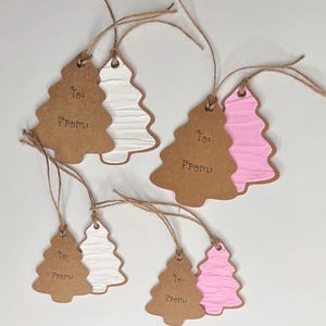 Op de afbeelding: Kerstboomvormige cadeaulabels. De labels zijn gemaakt van bruin karton met een getextureerd wit of roze boomontwerp. Elk label heeft de woorden "To:" en "From:" erop gedrukt en is geregen met touw.