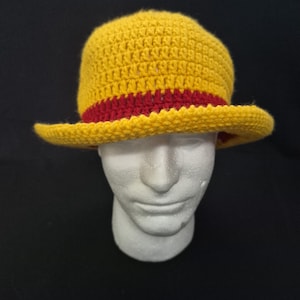 May include: A yellow crochet hat with a red band. The hat has a wide brim and a rounded crown. The hat is displayed on a white mannequin head against a black background. The hat appears to be handmade.