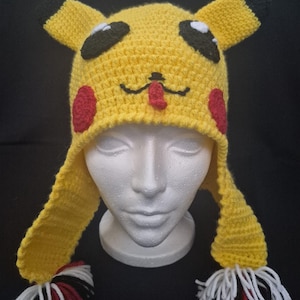 Puede incluir: Un gorro amarillo de ganchillo con orejas con puntas negras, ojos negros y blancos y círculos rojos en las mejillas. El gorro tiene solapas largas con borlas de hilo blanco, negro y rojo. El gorro está diseñado para parecerse al personaje Pikachu.