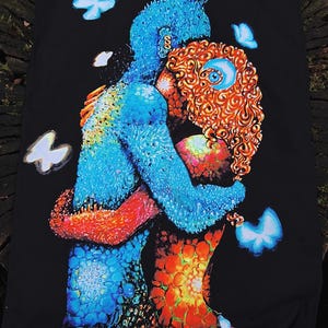 Op de afbeelding: Een zwarte wandtapijt met een levendige, abstracte afbeelding van twee figuren die elkaar omarmen. De figuren zijn weergegeven in tinten blauw, oranje en rood, met ingewikkelde patronen en details. Witte en blauwe vlinders zweven rond de figuren.