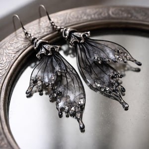 May include: A pair of silver butterfly wing earrings with detailed design. The earrings have delicate, translucent wings with a gradient effect, embellished with small, sparkling details. They are displayed on a decorative, silver-framed mirror.