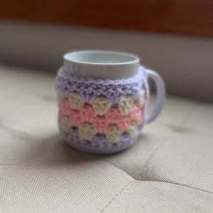 May include: A white ceramic mug with a crocheted cozy. The cozy features a granny square design in pastel colors: lavender, pink, and cream. The mug is on a neutral-toned surface.