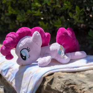 Könnte beinhalten: Ein hellrosa Plüschpferd mit einer leuchtend pinkfarbenen Mähne und Schweif. Das Pony hat große blaue Augen und liegt auf einer gemusterten Decke. Das Spielzeug liegt auf einer Steinoberfläche.