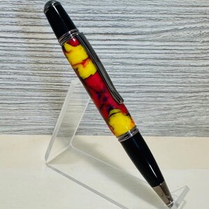 May include: A handcrafted pen with a vibrant design. The pen features a black top and bottom, a silver clip, and a colorful barrel with red, yellow, and purple swirls. The pen is displayed on a clear acrylic stand.