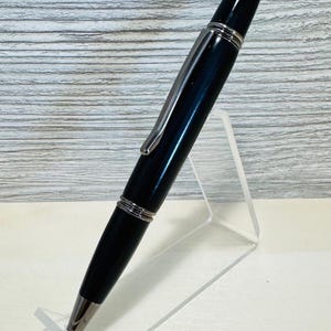 May include: A black and silver pen with a shiny finish, displayed on a clear acrylic stand. The pen has a silver clip and accents, with a silver tip and a rounded top. The pen is designed for writing.