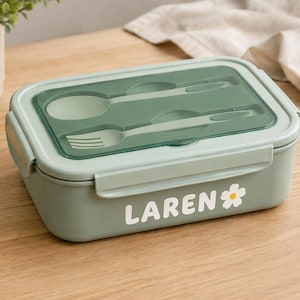 May include: A rectangular sage green lunch box with a translucent lid featuring a built-in spoon and fork. The side of the container displays the white text LAREN followed by a small white and yellow flower graphic, secured with side latches.