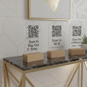 May include: Three arched clear acrylic signs with black printed QR codes and text. The first reads Scan to Play Our Song, the second Scan to Wi-Fi welcome our home, and the third Scan for a Memory Jane and Alex. Each sign sits in a rectangular wood base.