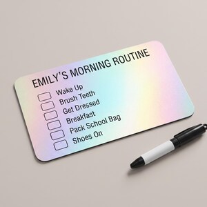 May include: A rectangular holographic checklist board with rounded corners featuring the text EMILY'S MORNING ROUTINE and a list of tasks including Wake Up, Brush Teeth, Get Dressed, Breakfast, Pack School Bag, and Shoes On, accompanied by a black marker.