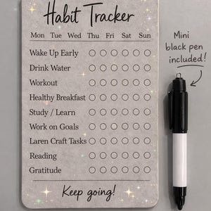 May include: A rectangular habit tracker board with a light grey speckled finish and sparkling accents. It features a list of daily tasks, columns for days of the week, and the text Habit Tracker and Keep going!. A mini black pen is included.