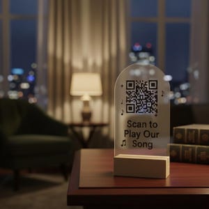 May include: A transparent acrylic sign with a QR code and the text "Scan to Play Our Song" is displayed on a wooden base. The sign is on a dark wood table with books, a lamp, and a green chair in the background.
