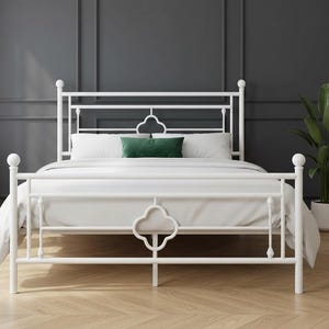 May include: A white metal bed frame with a decorative headboard and footboard. The bed is made up with white bedding and a green pillow. The room has a dark gray wall and a wooden floor.