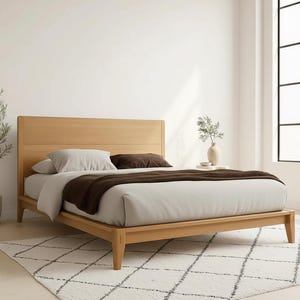 May include: A light wood bed frame with a headboard, featuring a light gray duvet, brown throw blanket, and pillows. The bed is on a white rug with a geometric pattern. A vase with greenery sits on a side table.
