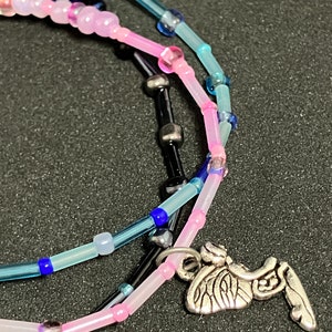 May include: A close-up of layered beaded necklaces in shades of pink, blue, and black. The necklaces feature small, colorful beads and a silver-toned charm shaped like a winged insect.